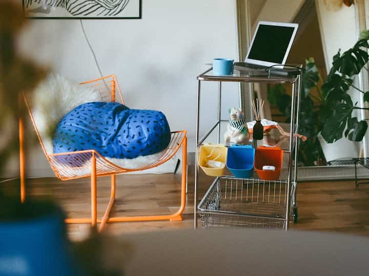 青いクッションとオレンジ色のワイヤーチェアがあるホームオフィス空間。金属製収納カートと植物で飾られた雰囲気のあるインテリア