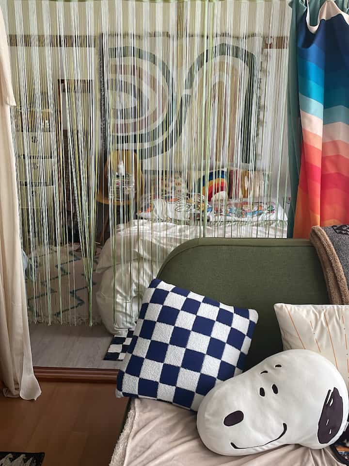 Natural modern small studio living room and bedroom featuring a green sofa, white bedding, rainbow blanket, and cushions with an open string curtain divider