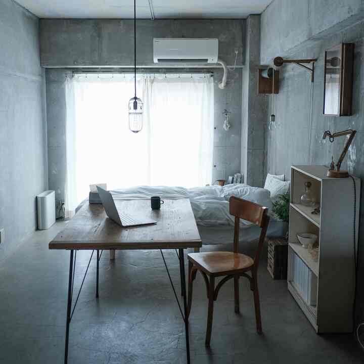 グレーのコンクリート壁と天井、白い寝具とカーテン、ナチュラルウッドのテーブルと椅子が配置されたシンプルな1人用ワンルームのリビング