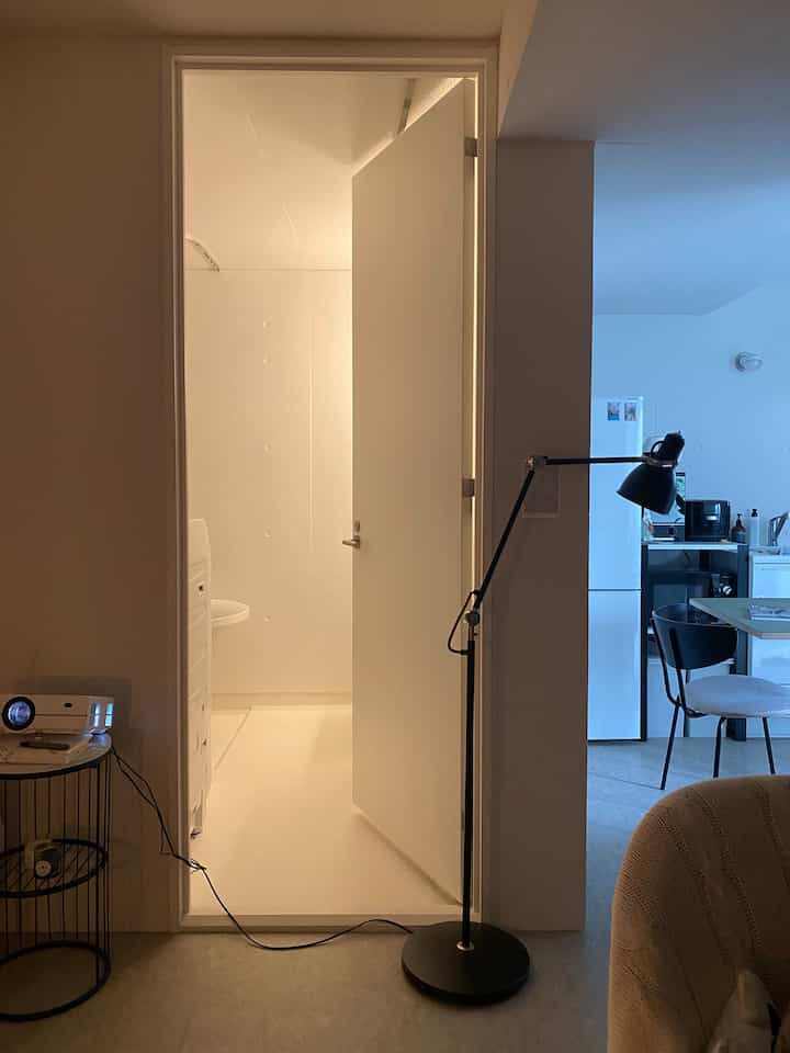 Modern studio apartment with bright white bathroom and kitchen, featuring a black floor lamp and simple furniture inside