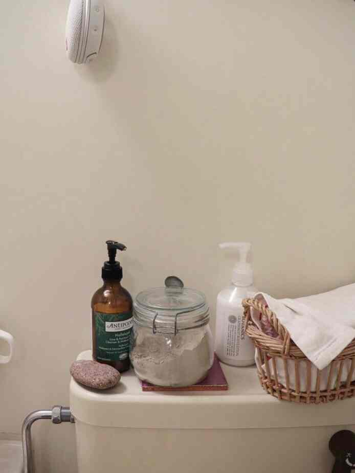 白とナチュラルな色調のバスルーム、トイレの上にクレンザーとタオルが置かれた落ち着いた雰囲気のインテリア