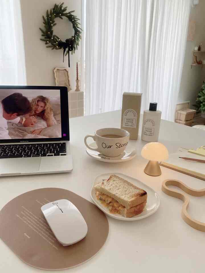 ホワイトトーンのホームオフィス、ノートパソコンとゴールドのミニテーブルランプ、コーヒーカップとサンドイッチが置かれた温かみのある作業空間