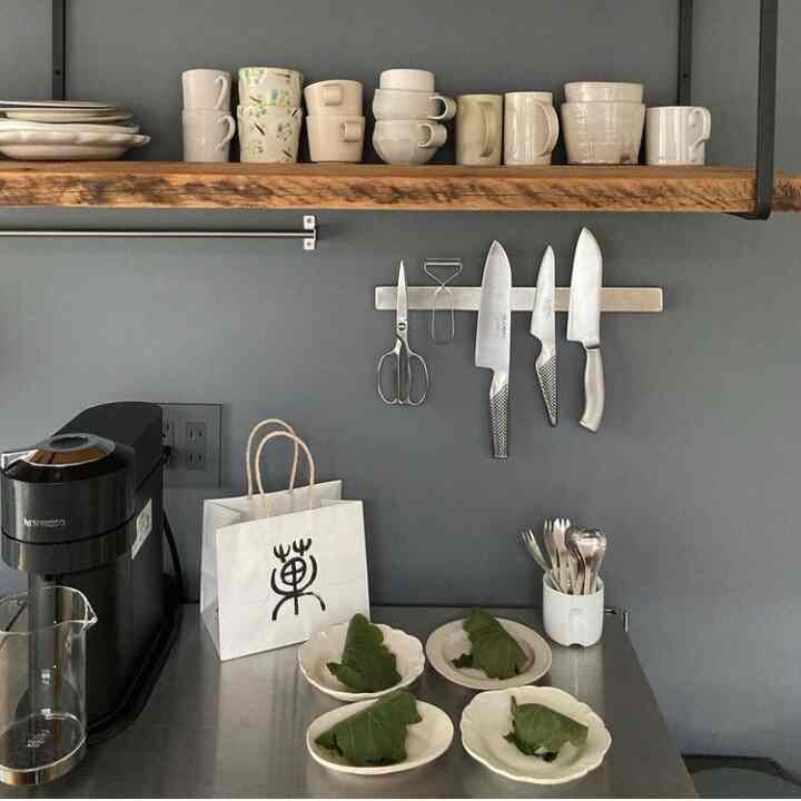 グレーの壁と白い食器が配されたシンプルなキッチン、ステンレスのカウンターと包丁が印象的な空間
