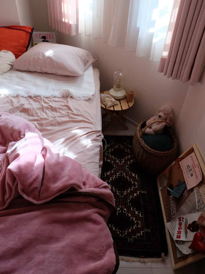 Cozy small bedroom with warm pink tones, featuring a plush bed, patterned rug, and curtains by the window with decorative elements