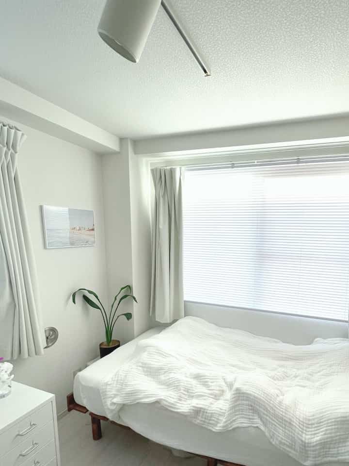 Bright and simple white-toned bedroom featuring a neatly made bed and blinds, creating a clean atmosphere