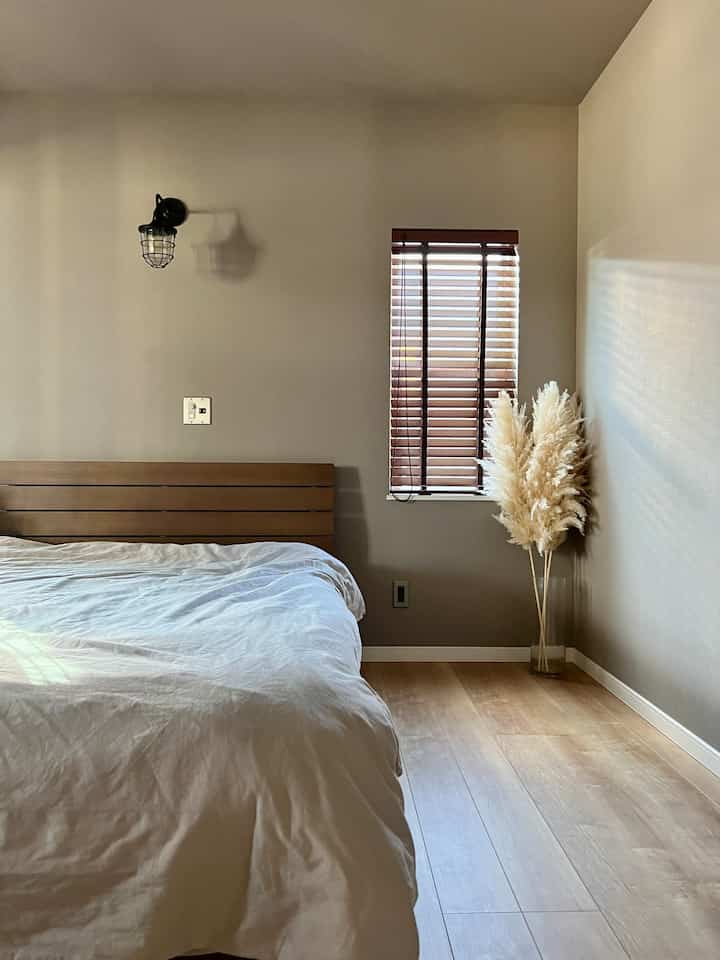 ベージュの壁とブラウンの木製ベッドが配置されたホテルライクな寝室、自然光とパンパスグラスが調和する落ち着いた空間