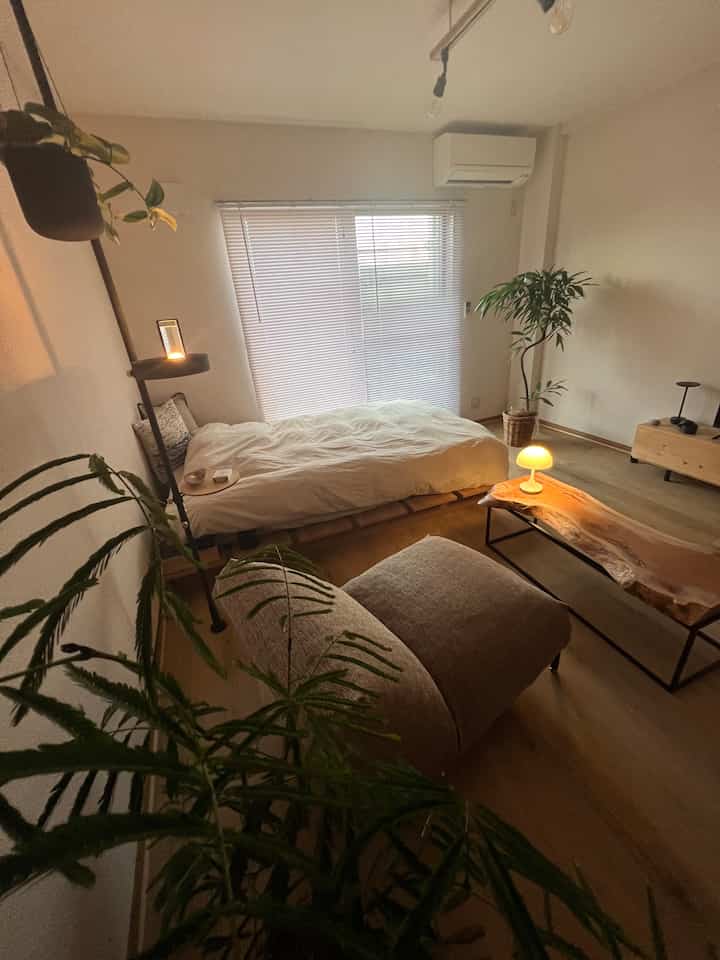 ナチュラルな木目調と白を基調としたコンパクトな寝室、オットマンと植物が配置された落ち着いた空間