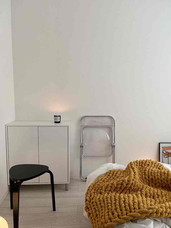 ホワイトトーンのミニマルなリビングスペース、キャビネットと透明な折りたたみ椅子、黒いスツールとマスタード色のスローが配置された落ち着いた雰囲気のインテリア