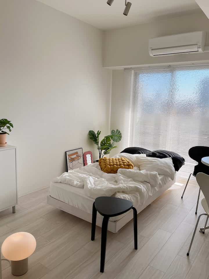 White-toned bedroom with a centrally placed bed, black stool in front, chairs near the window, and green plant creating a natural ambiance