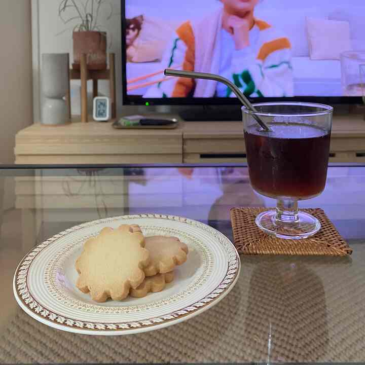 ナチュラルなトーンのリビング、ガラステーブルの上にラタンコースターと強化ガラスのグラス、クッキーが載った皿が置かれた暖かい雰囲気のインテリア