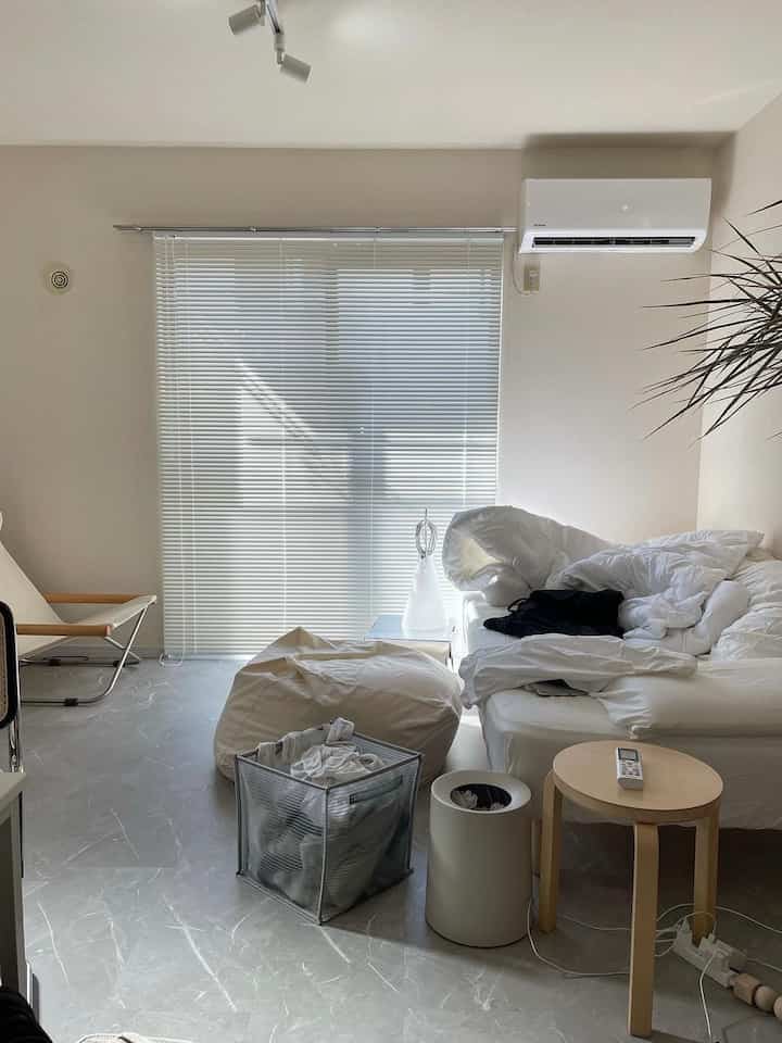 明るいベージュ色調の小さな寝室、白いソファベッドとビーンバッグ、光が差し込むブラインドがあるシンプルで自然な雰囲気の空間