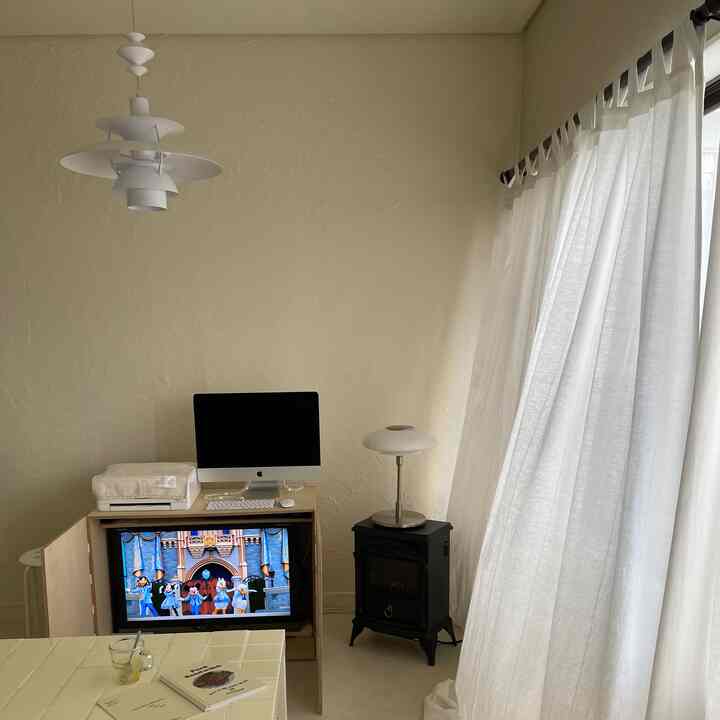 白を基調としたリビング、コンパクトな空間にテレビ台、パソコンデスク、電気暖炉、明るいカーテンが配置された雰囲気のあるインテリア