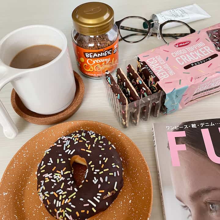 白いテーブルにホームカフェ用のマグカップとチョコレートドーナツ、ピンクのクラッカーパッケージと雑誌が置かれた雰囲気のある空間
