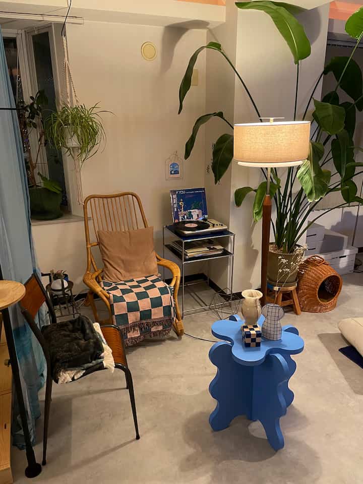 Natural-toned living room featuring a rattan armchair, blue side table, floor lamp, and large indoor plants creating a cozy atmosphere
