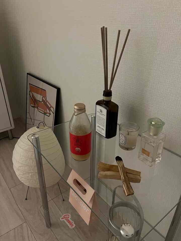 ベージュの床と透明なガラスラックが配置された寝室、ブラウンのディフューザーとキャンドルが置かれた雰囲気のある空間