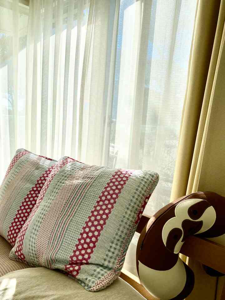 明るいナチュラルトーンのL字型寝室、木目調のベッドと枕、カーテン、そしてそばにナマケモノのぬいぐるみが配置された雰囲気のある空間