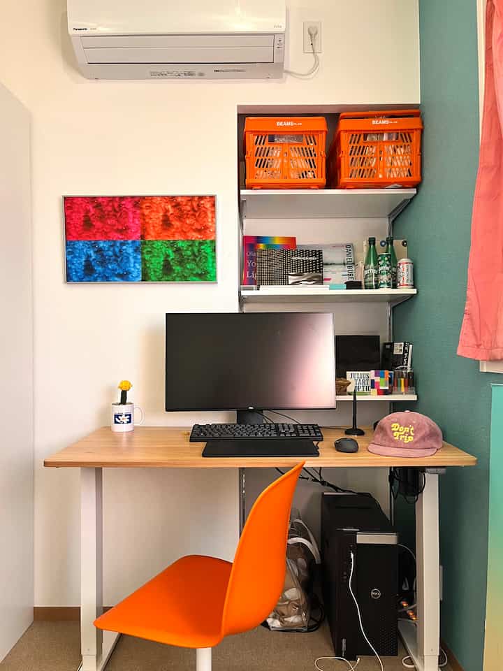 ホワイトの壁と木目調のデスクが配置された狭いホームオフィス、鮮やかなオレンジの椅子と収納用のオレンジのボックスが印象的なモダンなインテリア