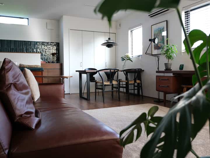 白い壁とブラウンのレザーソファが配されたナチュラルモダンなリビング、中央にはペンダントライトとダイニングチェアが置かれた落ち着いた空間