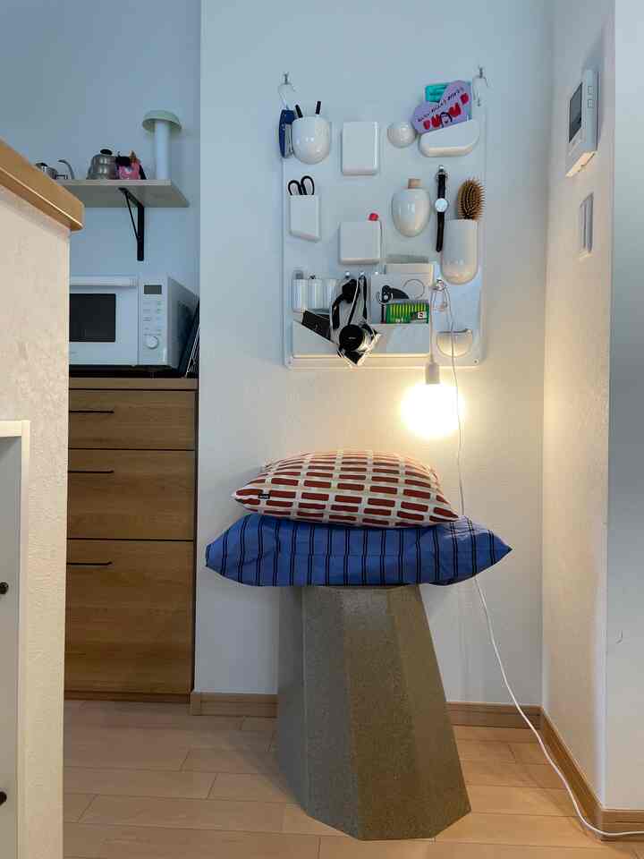 Bright white walls and wood-tone kitchen in a compact space featuring wall-mounted storage, cushions on a modern stool, and a glowing pendant light, creating a cozy atmosphere