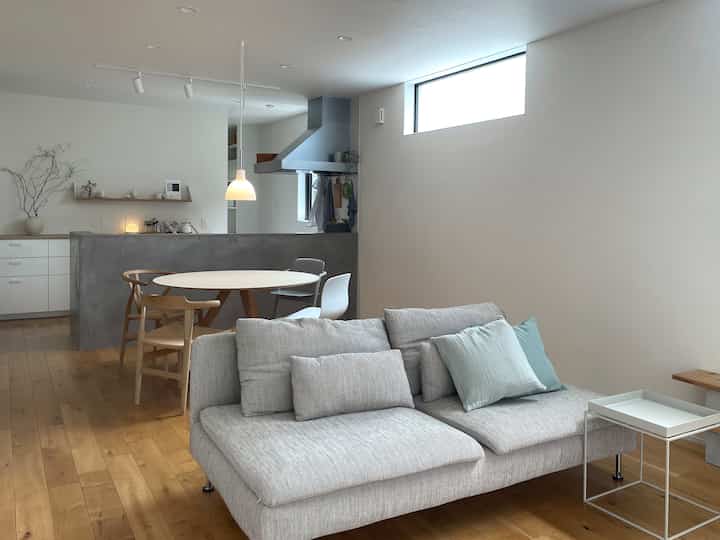 白い壁と木目調の床、グレーのソファと丸いダイニングテーブルが配置されたモダンなリビング兼キッチンスペース