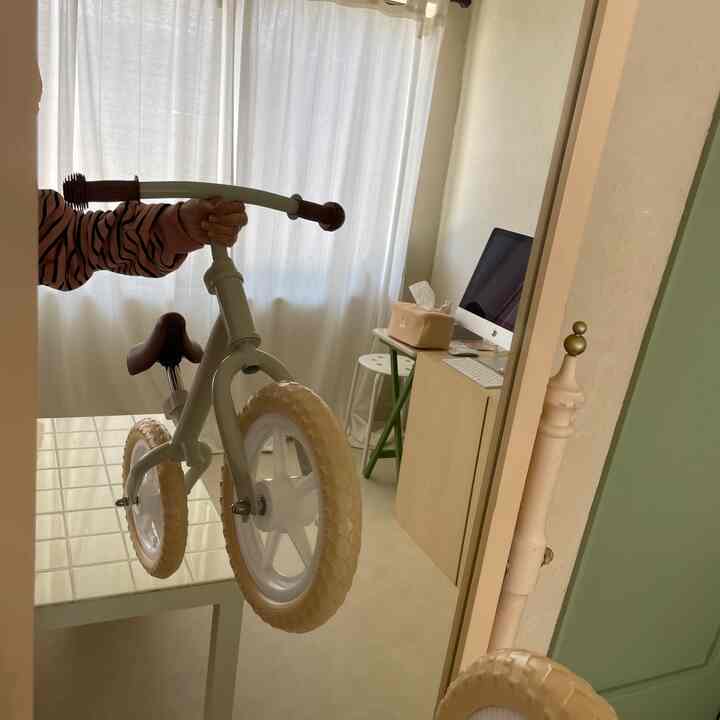 A white and beige toned kids' room featuring sheer curtains by the window, a child's balance bike, and a desk with computer setup