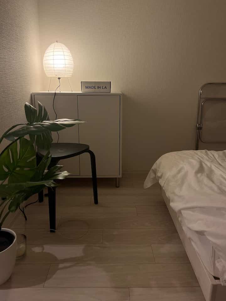 Natural tones and white dominate this bedroom featuring a white storage cabinet with a glowing lamp, a black stool, and a green plant creating a modern, minimal atmosphere