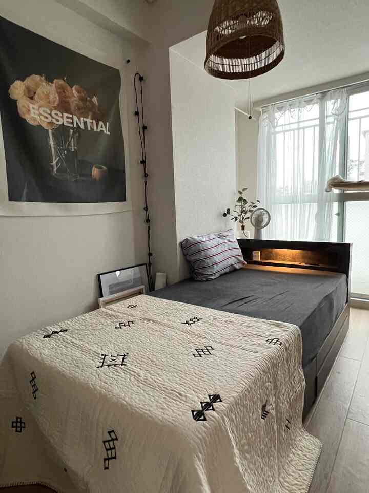 白い壁と木目調の床が特徴のナチュラルな小さな寝室、ヘッドボード付きのベッドとベージュのペンダントライトが配置された雰囲気のあるインテリア