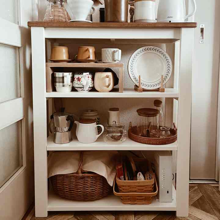 白と木目調のキッチン、整頓されたカップと皿が配置された収納棚が落ち着いたおうちカフェの雰囲気を演出しています。