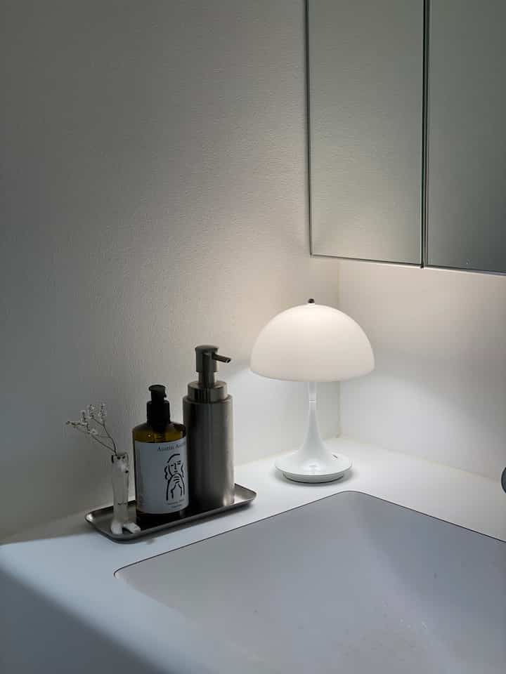 落ち着いたホワイトトーンのバスルーム洗面台空間、ステンレス製ディスペンサーとテーブルランプが配置されたシンプルなインテリア