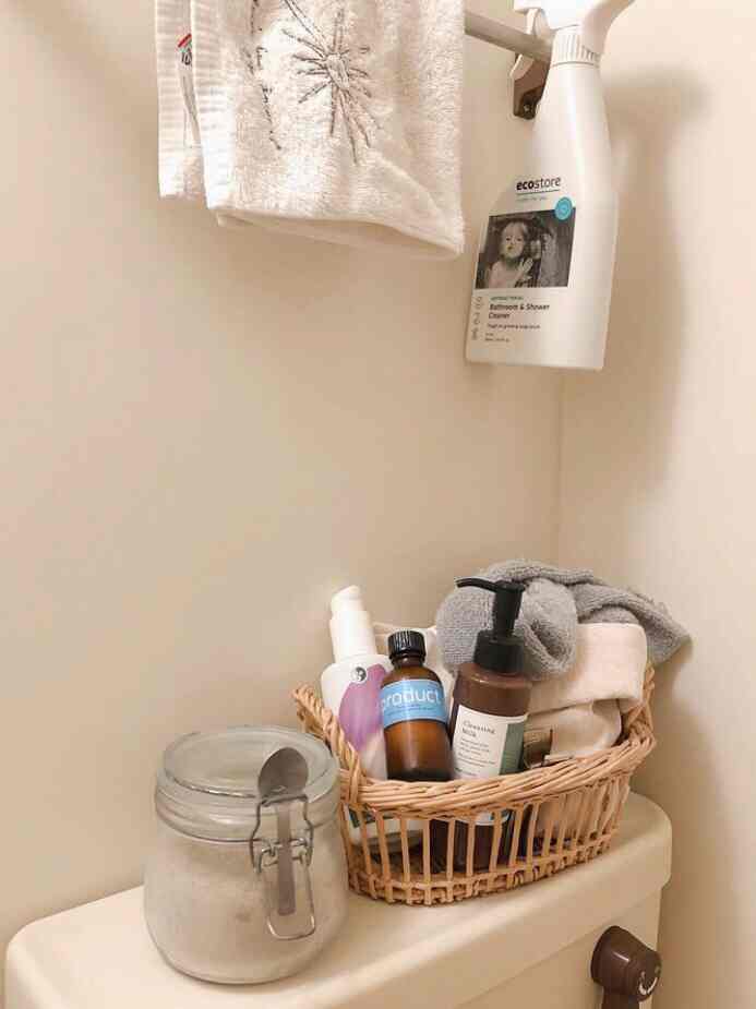 ベージュ色調のバスルーム、トイレの上にスッキリと収納されたバスクリーナーとタオルの置かれたバスケットが配置された雰囲気のあるインテリア