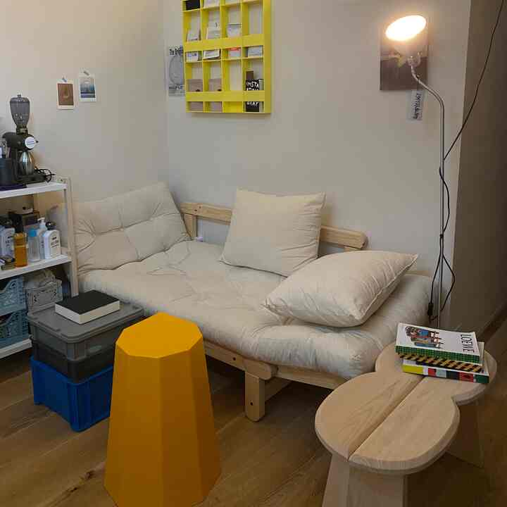 Natural-toned small living room featuring a white sofa bed, yellow stool, wooden table, and floor lamp creating a cozy atmosphere