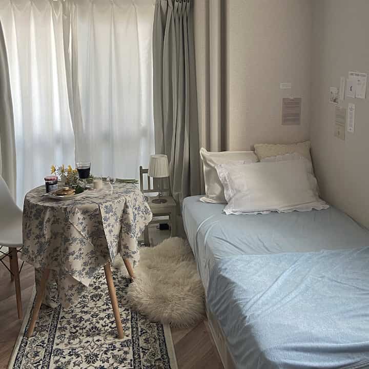 A beige and sky blue toned bedroom featuring a single bed, small dining table, and white curtains in a cozy single household space