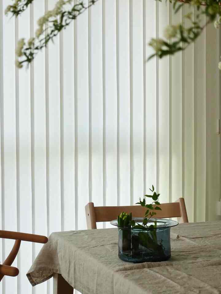 明るい白い縦型ブラインドを背景に、ベージュのリネンテーブルクロスと木製の椅子があるナチュラルなダイニング空間