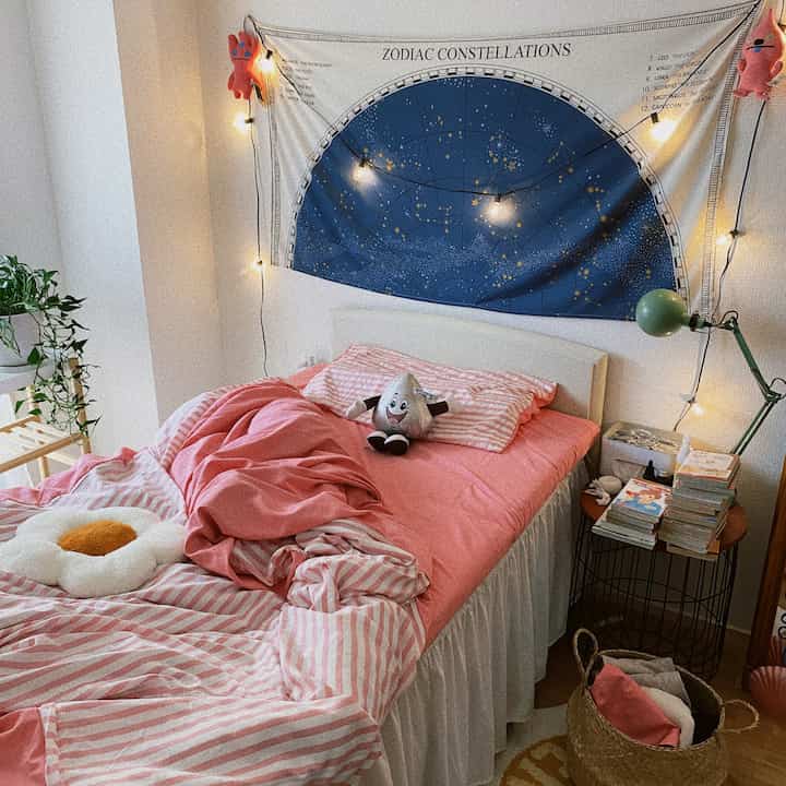 ピンクの寝具と星座のタペストリーを背景にした居心地の良い寝室、ストリングライトと本、植物が調和した雰囲気のあるインテリア