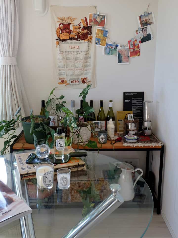 白い壁とカーテンのあるダイニング、透明ガラスのダイニングテーブルにキャンドルとルームスプレー、背後には植物とワインボトルのあるコンソールが配置された雰囲気のあるインテリア