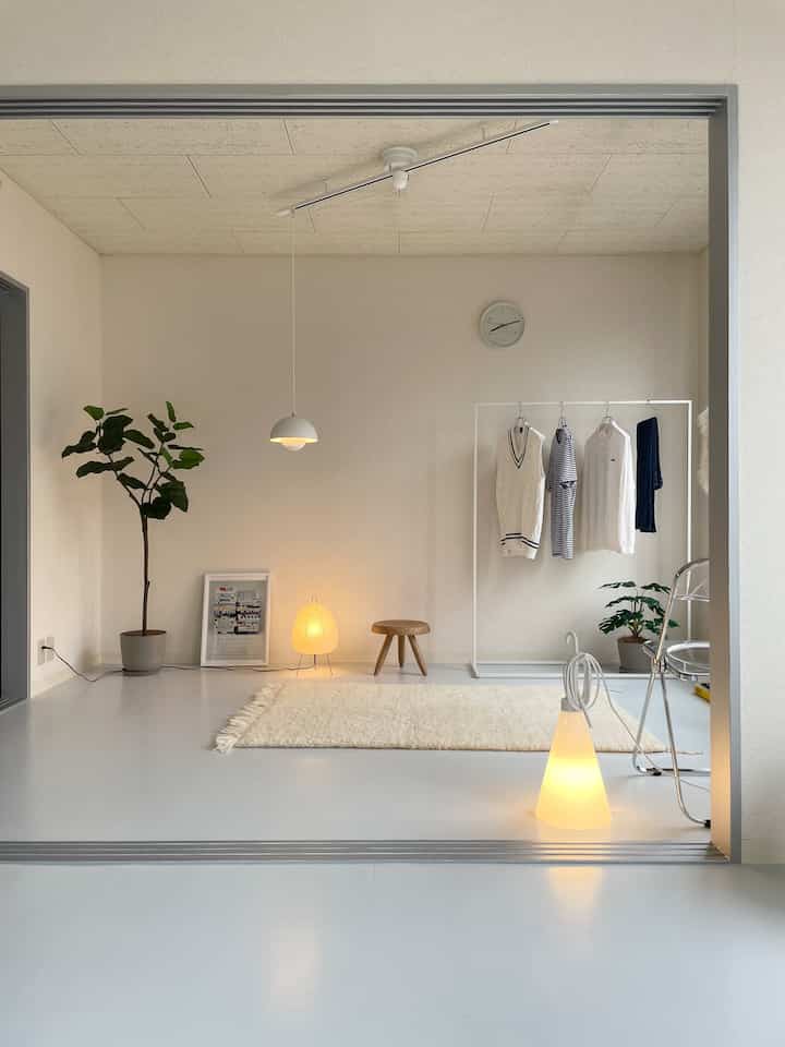 電球色の照明が温かみを演出する、シンプルなホワイトインテリアの部屋。収納用ハンガーと植物が配置された落ち着いた空間