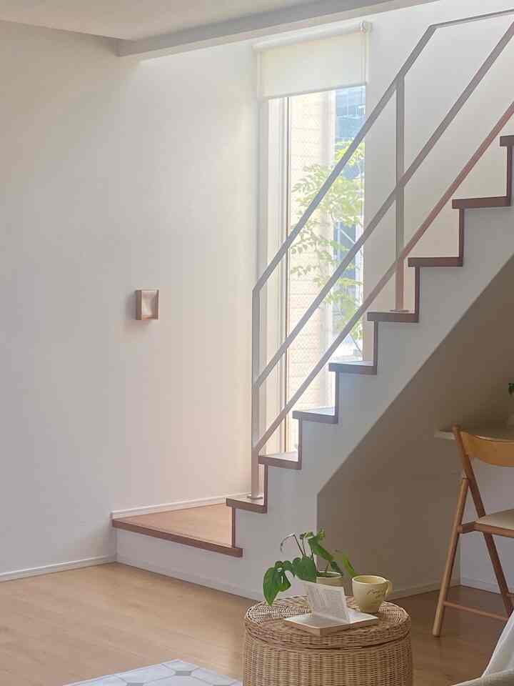 White walls and wood-tone stairs with a rattan side table and dining chair in a long and narrow living space