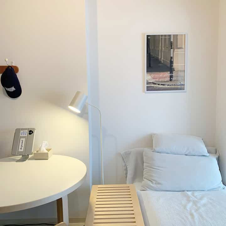 ナチュラルカラーのシンプルなリビング、白い丸テーブルとベージュのベッド、フロアランプが配置された落ち着いた雰囲気のインテリア