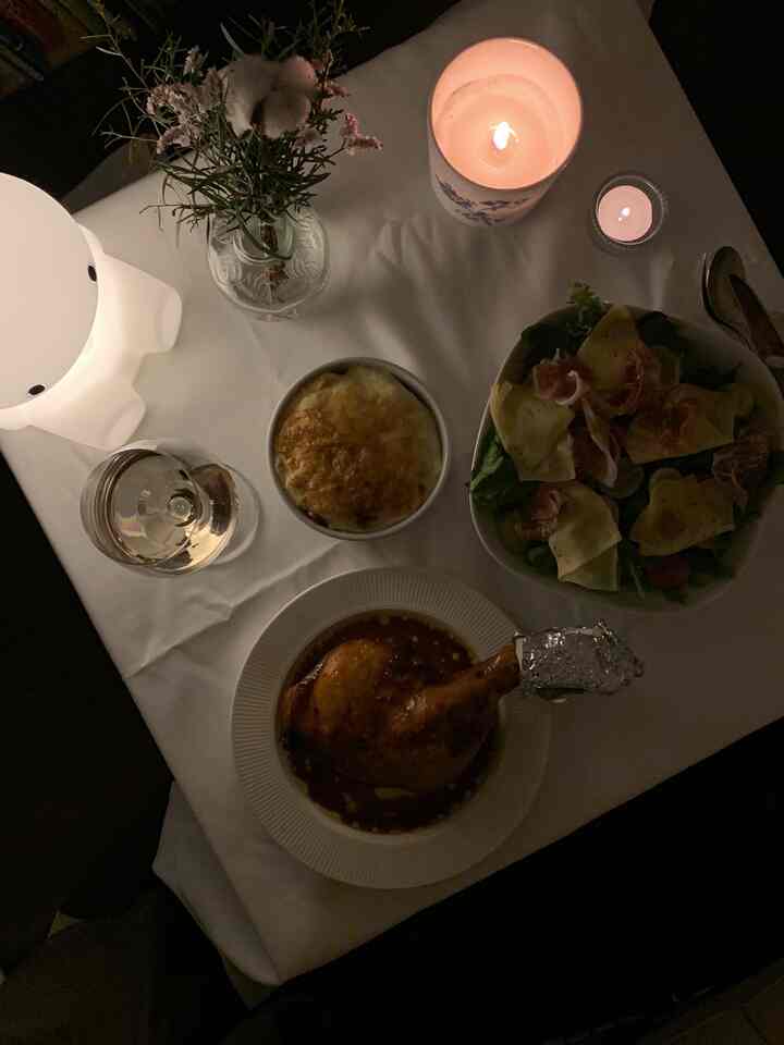 薄暗い照明の下、白いテーブルクロスの上にキャンドルと花瓶が置かれ、チキンレッグ、サラダ、グラタン料理が並んだダイニングのクリスマスディナーのセッティング