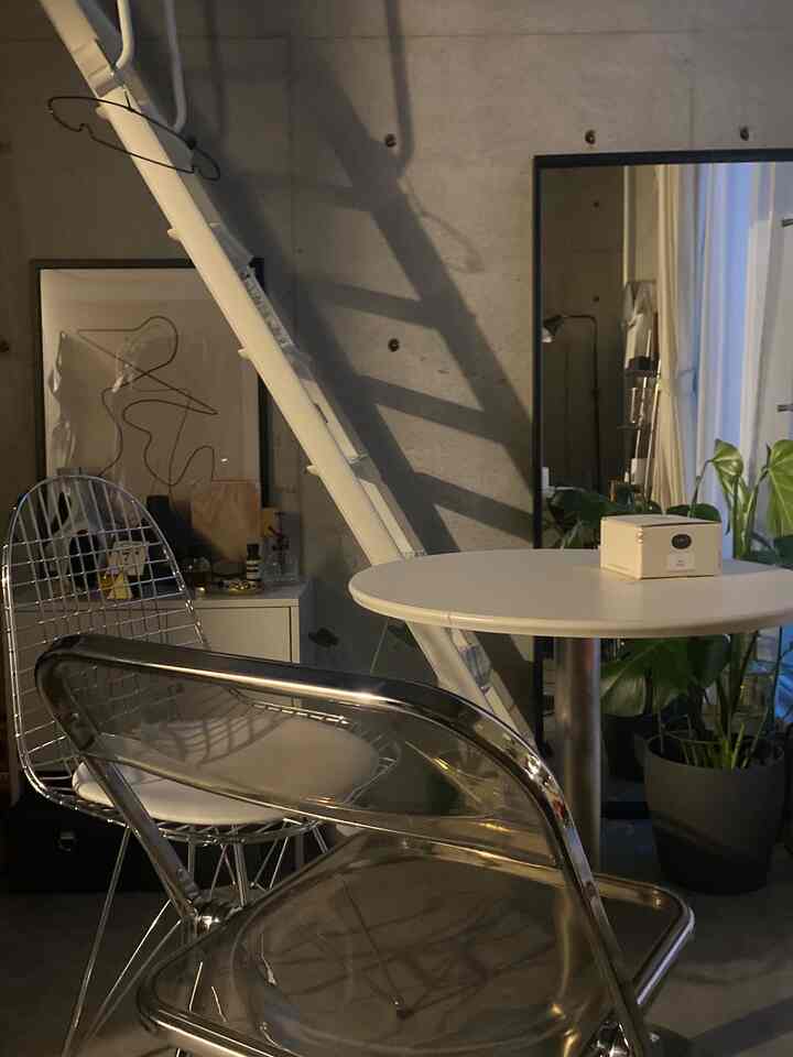 透明なチェアとシルバーのワイヤーチェアが配置されたミニマルでインダストリアルな狭い部屋、丸いテーブルと植物が調和した雰囲気のあるインテリア