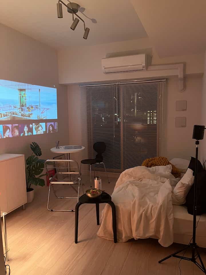 A cozy small living room in white and black tones featuring a sofa bed, transparent chair, blinds on window, and a projector screen on the wall