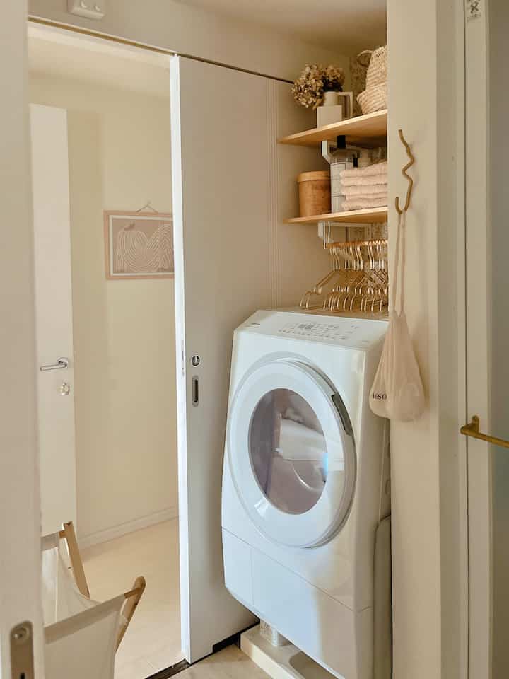 白とナチュラルウッドトーンの調和が取れた小さなワンルームのランドリールーム、ドラム式洗濯機と整頓された棚が配置されたミニマルな空間