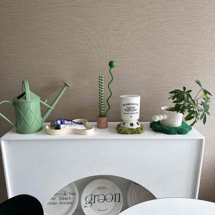 ベージュ色の壁と白い家具の上に緑のジョウロや植物、プレートが配置されたミニマルで自然な雰囲気のあるリビング