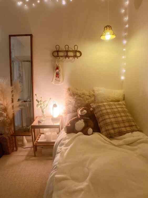 Beige and white toned bedroom featuring cozy bedding, cushions, table lamp, and string lights creating a warm atmosphere