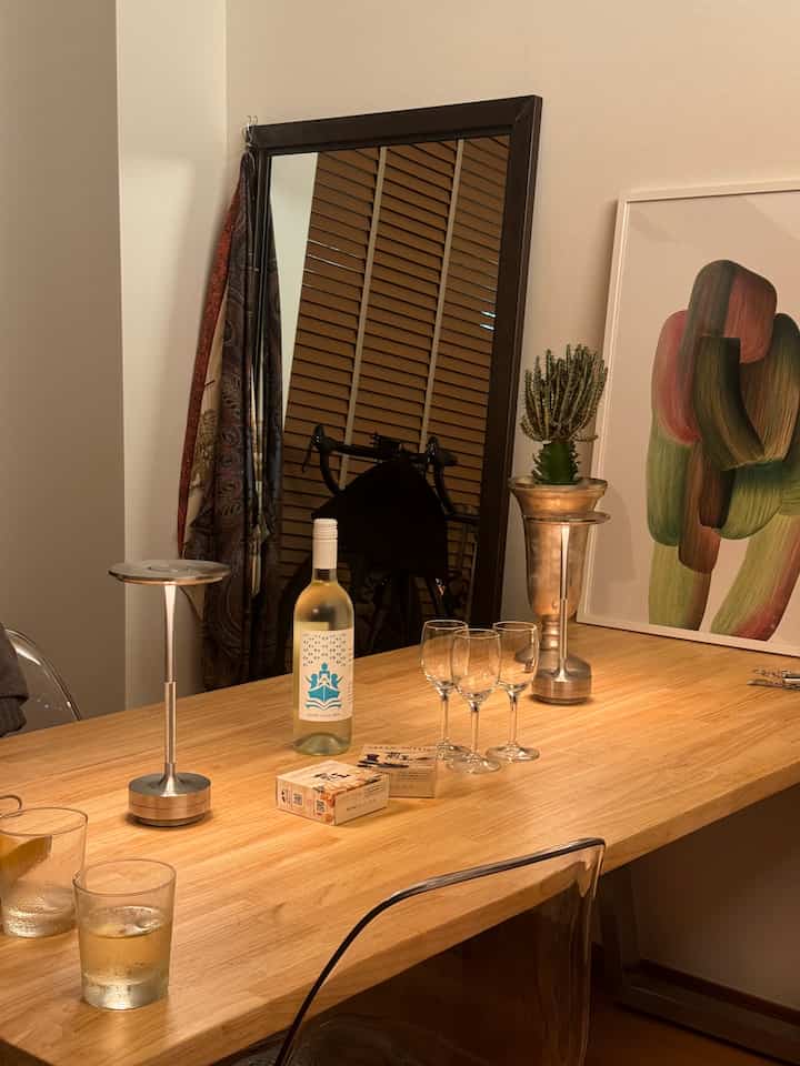 ナチュラルな色調のダイニングルーム、木製のダイニングテーブルとアルミニウム製のテーブルランプ、ワイングラスとポスターが配置された温かみのあるインテリア