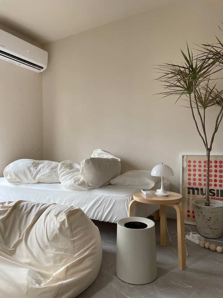Natural modern tone bedroom featuring white bed, stool, wood nightstand, plant, and poster creating a cozy atmosphere