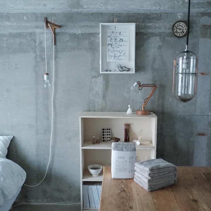 ナチュラルカラーとグレーのトーンが特徴の寝室、木製テーブルの上に整然と積まれたグレーのタオルがあるミニマルなインテリア