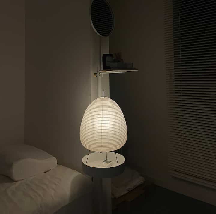 柔らかい白色の光が灯る寝室、中央に和紙のフロアランプと小さな棚が配置されたシンプルなインテリア