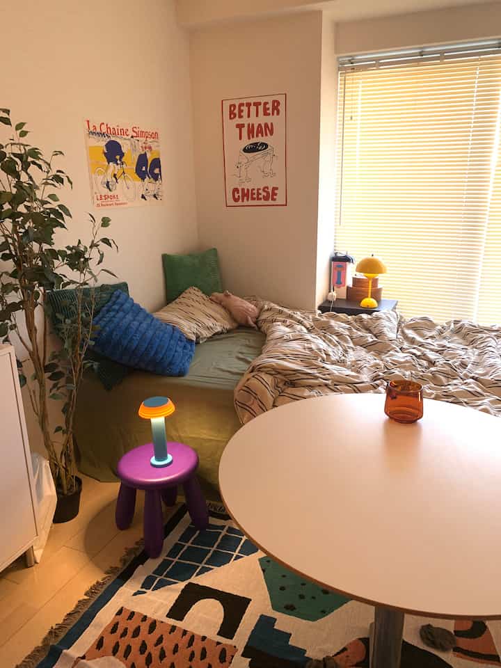 Cozy small bedroom featuring a light gray table, green bedding, purple stool with lamps, and a plant