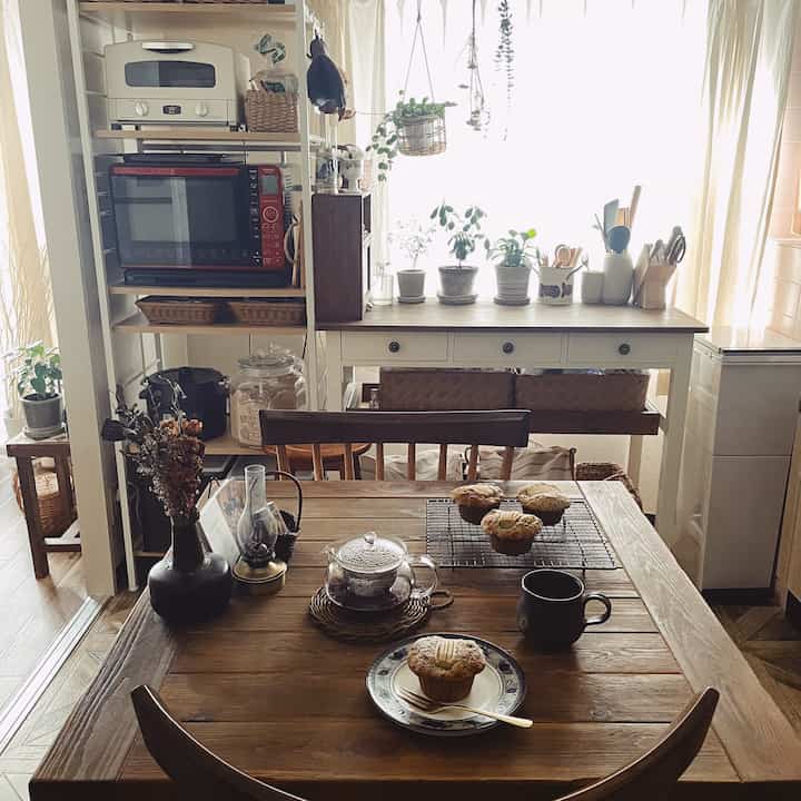 木目調でナチュラルなキッチン兼ダイニング、中央に食卓と椅子、食卓にはマフィンとティーセットが配された雰囲気ある空間